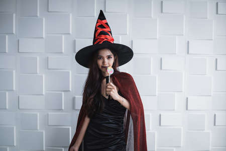 Young Asian woman in costume witch and hold lollipop on white wall of Halloween concept. Portrait of teen woman dressed up as witch for celebrate Halloween festival.の写真素材