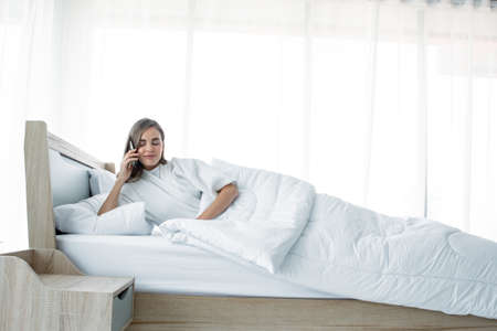 Woman talking smartphone on bed with a smiling face after waking up in the morning.の写真素材