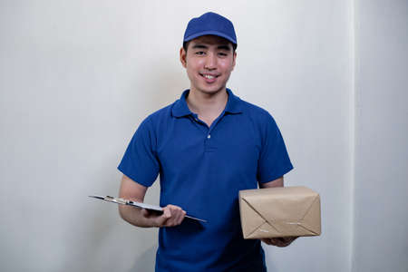 Happy young delivery man in blue dress with parcel post box and document on white background with...