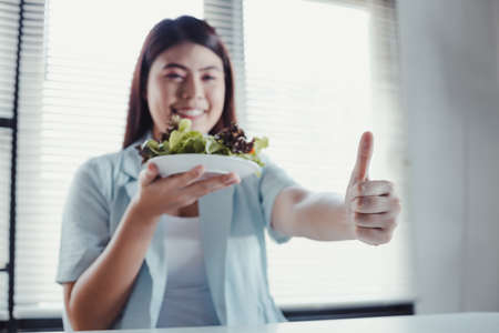 Asian woman thumbs up and holding dish vegetable for healthy diet with smile. Beautiful smiling woman eating fresh organic vegetarian salad. Young woman eating healthy salad in diet concept.の写真素材