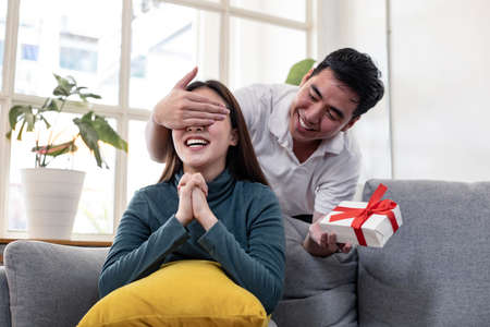 Young man closed his girlfriend's eyes to surprise with a gift box held on the occasion of the anniversary of love. Happy man surprises his girlfriend with gift box at home with smile.の写真素材