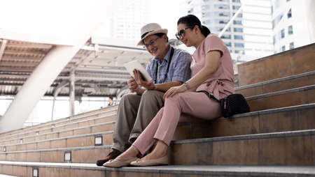 Senior Asian couples sit on stairs, plan, find travel information via tablet with smile. Senior couple travel concept. Aunt and uncle sitting on stairs with playing tablet while traveling for tourism.の写真素材