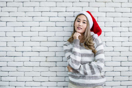 Portrait cute Asian young woman in costume Christmas and wear red santa claus hat with smile on white brick background and copy space. Happy of teen woman in theme Christmas.の写真素材