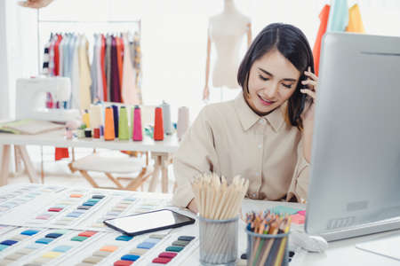 Beautiful Asian woman designer, talking on the phone with the customer and taking note of the work details in order to receive the tailor order for the customer with smile. Startup designer concept.の写真素材