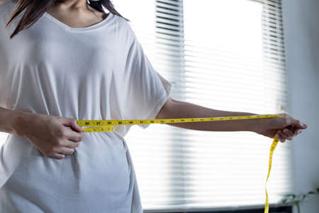 Slim woman measuring her waist by measure tape after diet. Girl with centimeter. Young woman with perfect waist with measuring tape. Hands measuring with tape at abdomen.