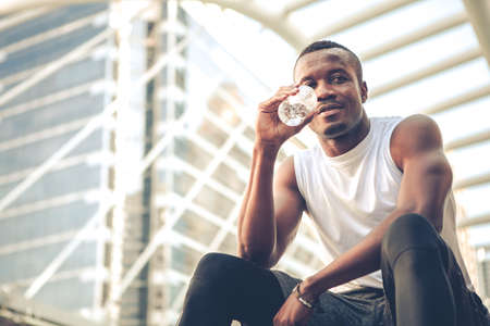 Portrait runner young African American man sitting drinking water with smile on the stairs while...