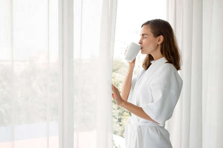 Beautiful Young Caucasian woman in pajamas holding a glass of white coffee ready to drink by the window in the bedroom at home after wake up in the morning. Woman standing drinking coffee by window.の写真素材