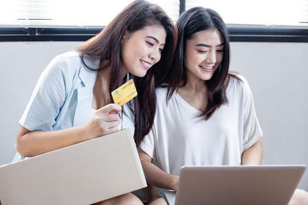 Two friends shopping online with credit card and laptop at home. Two happy young Asian woman holding laptop computer with credit card while sitting in the home.の写真素材