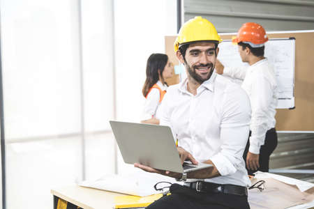 Young engineer is working and holding a laptop, with others engineers planning on the board in the...