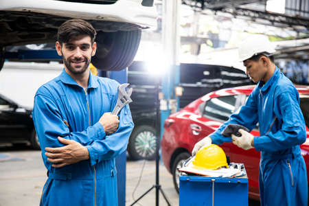 Confident handsome smiling mechanic in uniform standing with his arms crossed and smiling while workmate working on background. Car mechanic working at automotive service center.の写真素材