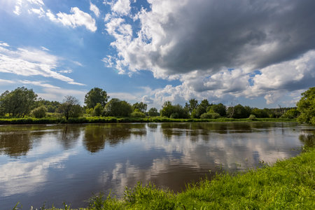 A large cloud is reflected in the river. Rural landscape. Bright green grass on the river bank....