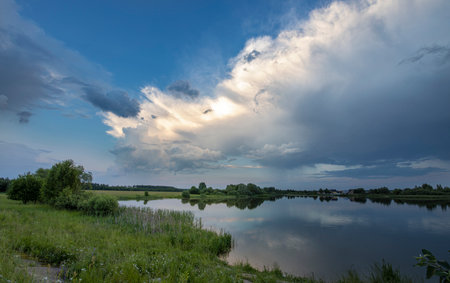 Rain clouds lit by the sun over the river after rain. bright green grass on the shore. A dramatic cloud in the evening sky, illuminated by the sun.の写真素材