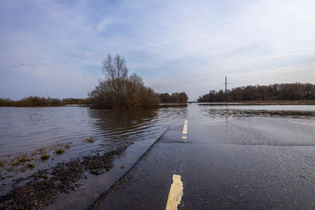 The road is under water due to flooding. The river overflowed its banks and flooded the road. Asphalted road between forest and field, flooding the road in a spring rural landscape.の写真素材