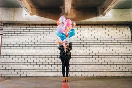 Girl with the bunch of multicolored balloons standing in front of the white brick wall in the parkingの写真素材