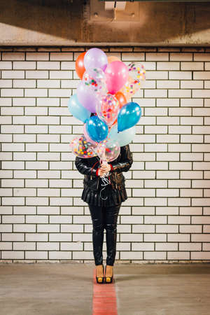 Girl with the bunch of multicolored balloons standing in front of the white brick wall in the parkingの写真素材