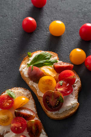 Flat lay with two bruschetta with curd cheese, cherry tomatoes and basil on a dark concrete...