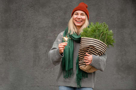 An attractive blonde holds a Christmas fire and fir branches in her hands and laughs against the background of a gray wall. Christmas mood. The holiday comes to usの写真素材