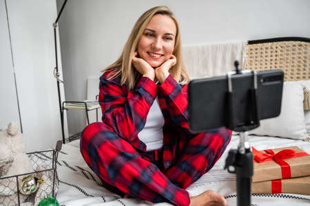 A young beautiful blonde is video chatting with her family and friends for the celebration of Christmas and New Year, sitting in a cozy bed, in traditional blue and red pajamas, surrounded by gifts and holiday decorations. Video callの写真素材
