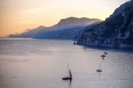 The rocky shoreline near Positano. A beautiful sunset light illuminates the quiet scene, a ray of light pierces the top of the mountains and reaches a flat sea where some boats are moored or sailing.の写真素材