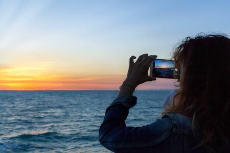 A girl photographs the sunset over the sea with her cell phone.の写真素材