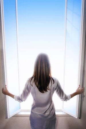 A woman with long hair and shrugging as she opens the window doors to let the light into the room. Space for text.の写真素材