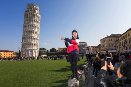 Pisa, Italy - February 11, 2018: Asian girl poses for a picture with the famous Tower of Pisa in the background, in Piazza dei Miracoli crowded with tourists.のeditorial素材