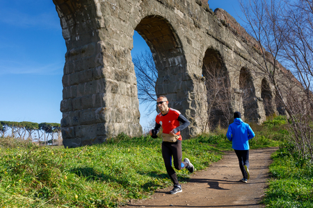Rome, Italy - January 26, 2019: two men run and train on the dirt path, in the public park. In the background the imposing ruins of the ancient Roman aqueduct.のeditorial素材