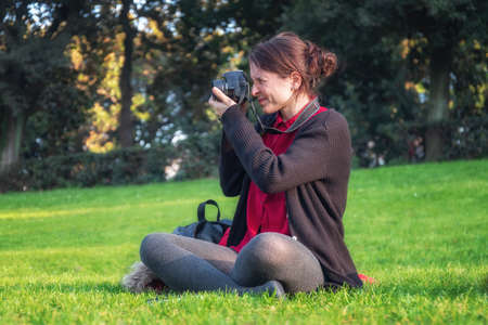 Girl sitting in the grass of the public park, photographing the surrounding landscape with her...