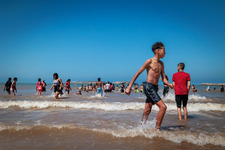 Essaouira, Morocco - August 3, 2023: Group of children jump into the sea to swim, only the girls wear very long dresses or costumes, as dictated by their religion.のeditorial素材