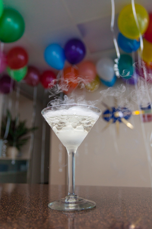 A glass with a transparent cocktail and thick smoke on the bar counter. Behind the blurred background is a room with balloons at the ceiling. Party, celebration, corporateの写真素材