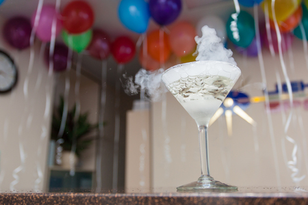 A glass with a transparent cocktail and thick smoke on the bar counter. Behind the blurred background is a room with balloons at the ceiling. Party, celebration, corporateの写真素材