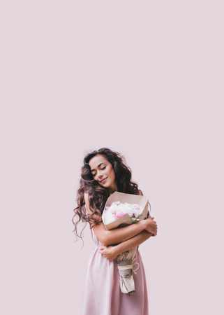 Young beautiful woman with long curly hair in a pink dress with a big bouquet of peonies on a pink backgroundの写真素材