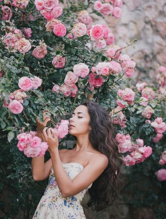 Young beautiful woman with long healthy curly hair wearing pink vintage dress and posing in garden with lots of wild roses.の写真素材
