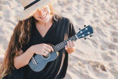 Beautiful young woman with long curly healthy hair wearing trendy straw hat playing ukulele at the beach.の写真素材