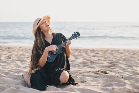 Beautiful young woman with long curly healthy hair wearing trendy straw hat playing ukulele at the beach.の写真素材