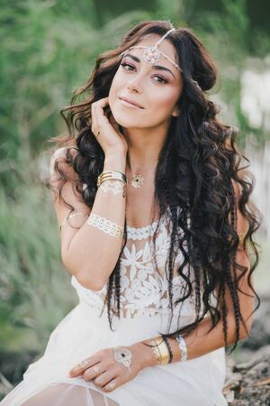 Beautiful young woman with long curly hair dressed in boho style dress posing near lake.の写真素材