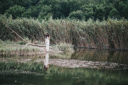 Beautiful young woman with long curly hair dressed in boho style dress posing near lake.の写真素材
