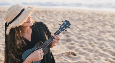 Beautiful young woman with long curly healthy hair wearing trendy straw hat playing ukulele at the beach.の写真素材