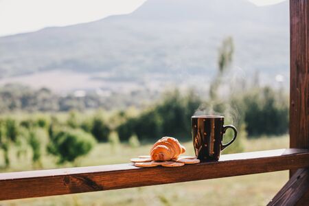Cup of hot tea and fresh croissant standing on cozy balcony with beautiful mountains view.の写真素材