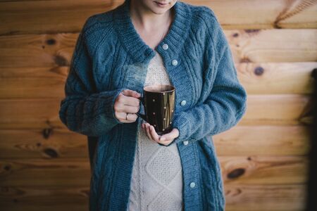 Woman in warm woolen knitted sweater witn cup of hot tea standing on cozy wooden balcony of country house.の写真素材