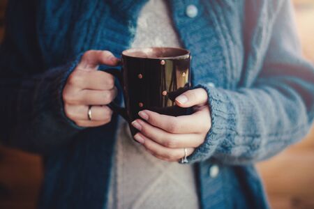 Woman in warm woolen knitted sweater witn cup of hot tea standing on cozy wooden balcony of country house.の写真素材