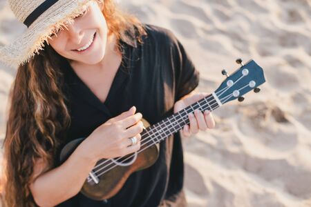 Beautiful young woman with long curly healthy hair wearing trendy straw hat playing ukulele at the beach.の写真素材