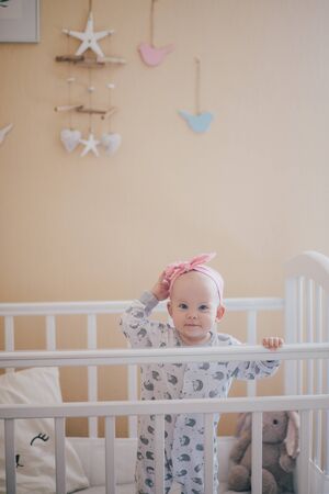 Adorable baby girl wearing cute pajamas standing in her white bed in a nursary room with beige walls.の写真素材