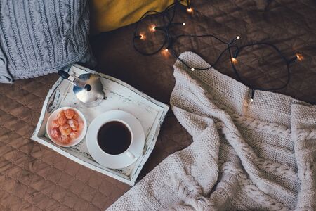 Still life with hot coffee and kumquat on wooden tray standing on bed with cozy lights on the background.の写真素材