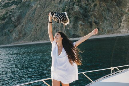 Young beautiful happy woman with long curly hair in hat and beach clothes posing on a yacht with sea and mountains on the background.の写真素材