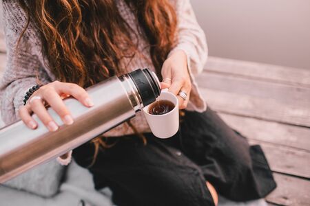 Happy woman in cozy knitted sweater pouring tea  in cup. She is sitting near lake in autumn forest.の写真素材