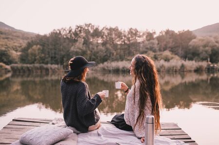Two female friends in knitted warm sweaters having picnic near lake with autumn forest and lake on...