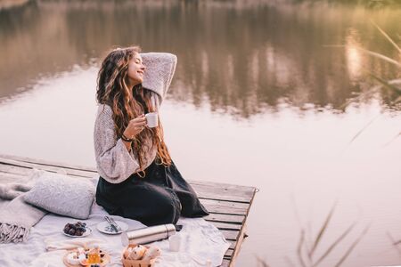 Happy pregnant woman in cozy knitted sweater drinking tea  . She is sitting near lake in autumn forest.の写真素材