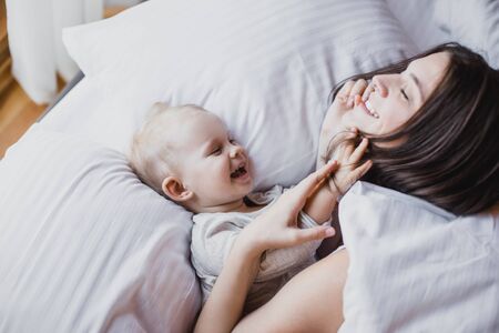 Happy young mother and her baby playing in bed with white linens. Happy family concept.の写真素材