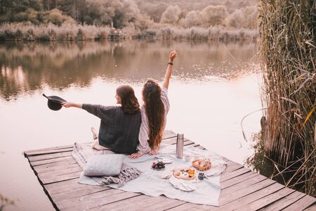 Two female friends in knitted warm sweaters having picnic near lake with autumn forest and lake on the background. Cozy fall atmosphere.の写真素材